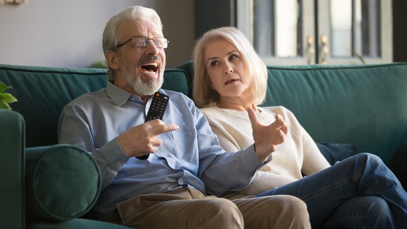Angry father-in-law watching TV with his wife who looks like she regrets marrying him in the first place