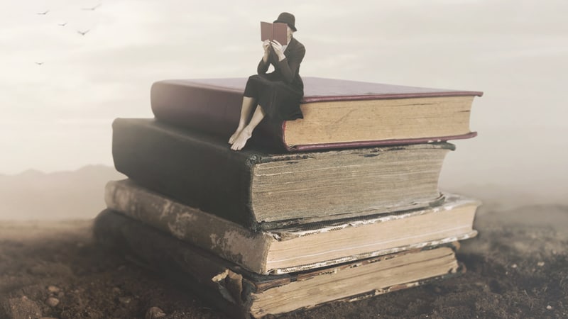 Surreal picture of a woman reading on top of a stack of old books. For Book recommendations for summertime reading