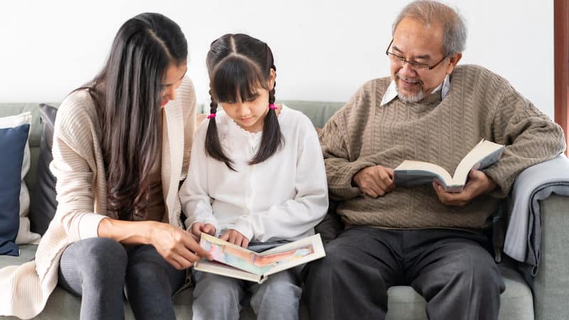 Multigenerational family - Asian mom, daughter, and granddad reading on family sofa, for