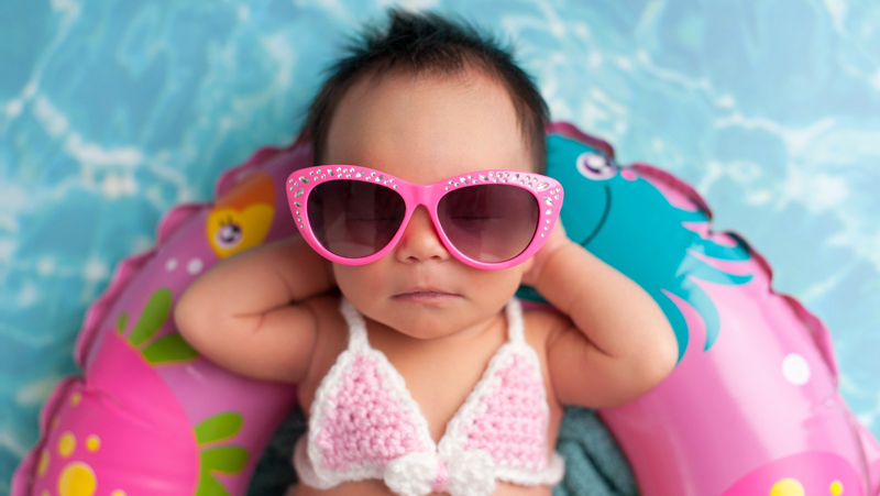 Newborn baby girl wearing pink crocheted bikini top and sunglasses, on a pool float Photo Katrina Trninich Dreamstime. For article on 2021 baby-naming inspirations