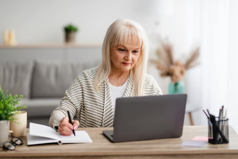 Woman at a laptop computer, with a notebook besides the laptop, possibly working on a puzzle