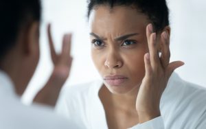 Black woman looking in a mirror concerned about her skin. For article on the skin-stress connection Image
