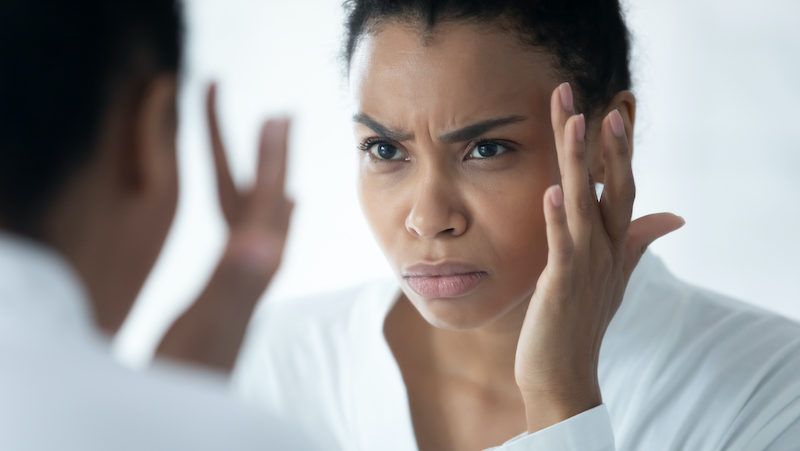 Black woman looking in a mirror concerned about her skin. For article on the skin-stress connection