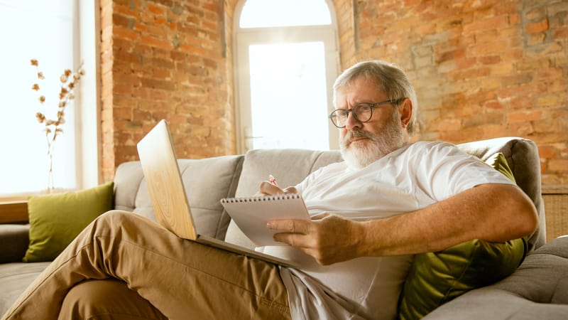 Man sitting on sofa with paper and laptop, possibly doing a puzzle. Image by volodymyr melnyk dreamstime