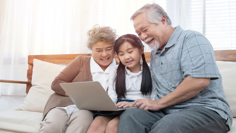 grandparents granddaughter looking together at a laptop, possibly doing a puzzle, sitting on a sofa. Photo by Champaiporn Kitina Dreamstime