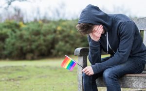 sad teenage boy holding pride flag photo by Ben Gingell Dreamstime. After he wears a gown to his prom, Grandma worries about the safety of her gay grandson. Is he going through a phase? See “Ask Amy” says. Image