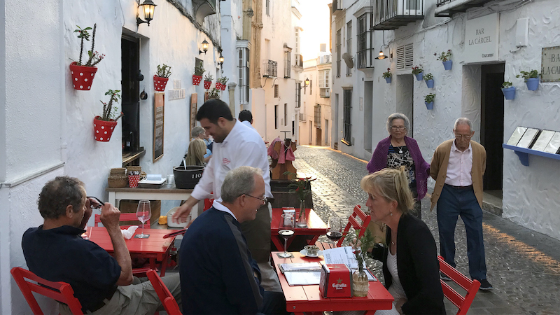 A cobbled street in Arcos serves as an alfresco dining spot. In Spain’s Andalucía region, stroll the streets of Arcos de la Frontera, sample market foods, and watch bull-fighting on TV at a bar. Image
