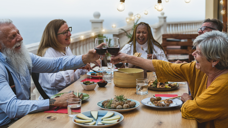 multiracial senior friends at a table outside, with food and wine, laughing. Alessandro Biascioli Dreamstime. Boomer reader Kate M. Carey bemoans little aches and pains, remedies, and early evenings that infiltrate life with her husband and friends. Image