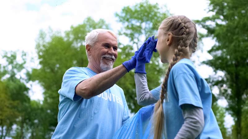 Senior man volunteering at a park clean-up event, high-fiving a young girl who is also volunteering. Looking for wise ways to use your retirement? Consider these 5 ways to give back and make your community and the world a better place.