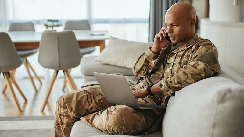Soldier sitting and talking on a phone while also looking at a laptop for resources, such as resources are available to help veterans managing IBD (inflammatory bowel disease), such as Crohn’s disease and ulcerative colitis. Image courtesy of Getty Images via Family Features.