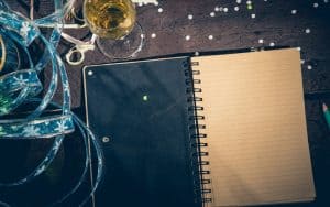 Ribbons, Champagne, and confetti beside a journal for writing next year's goals. Looking for new goals for the upcoming year? You can adopt these tongue-in-cheek New Year’s resolutions as your own, laughing all the way. Image