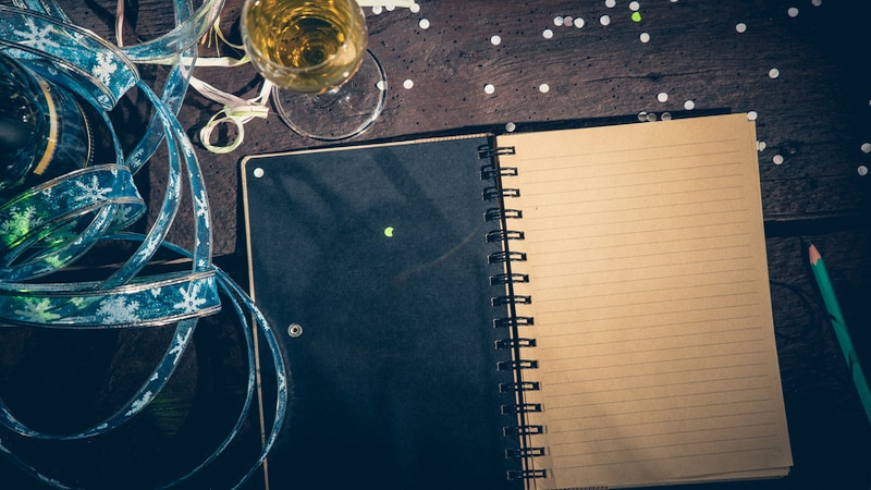 Ribbons, Champagne, and confetti beside a journal for writing next year's goals. Looking for new goals for the upcoming year? You can adopt these tongue-in-cheek New Year’s resolutions as your own, laughing all the way.