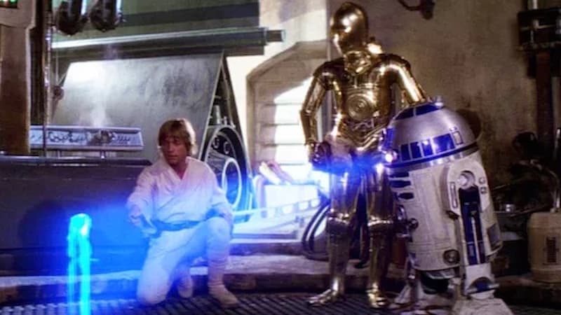 Luke Skywalker, C-3PO, and R2-D2, looking at a hologram in the movie "Star Wars." For article on five movies that predicted the future and what they predicted.