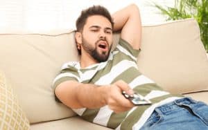 lazy young adult man on sofa using remote. A mother recognizes that she and her husband are enabling their adult son, but her husband doesn't agree. See what “Ask Amy” advises. Image