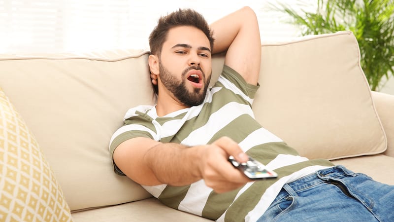 lazy young adult man on sofa using remote. A mother recognizes that she and her husband are enabling their adult son, but her husband doesn't agree. See what “Ask Amy” advises.