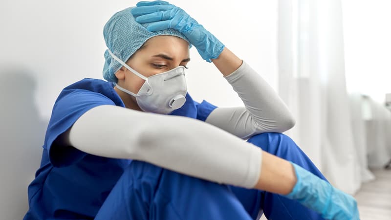 a sad nurse or doctor wearing PPE and sitting on the floor of a hospital. Image by Syda Productions. A nurse is tired of hearing people complain about working from home. See what “Ask Amy” says about post-pandemic problems and perspectives.