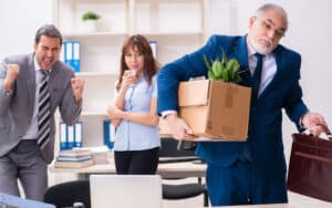 Man leaving office upon retirement, with box and briefcase, with younger coworkers laughing behind his back. What is retirement etiquette when retiring from a company that disrespects you? Image