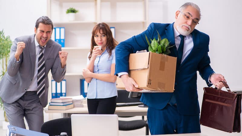 Man leaving office upon retirement, with box and briefcase, with younger coworkers laughing behind his back. What is retirement etiquette when retiring from a company that disrespects you? Image