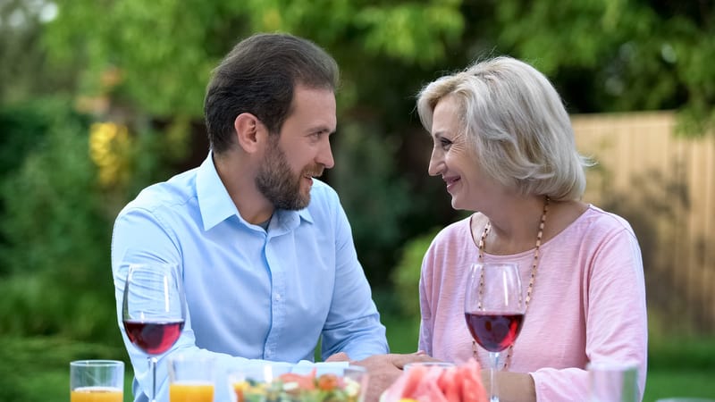 A middle-aged man and older woman smiling at each other over outdoor dinner and glasses of red wine. Image by motortion. For article, Her son-in-law uses her granddaughter as a pawn, refusing to let them get together. What does “Ask Amy” advise for this controlling man?