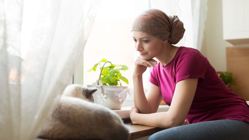 young woman with cancer head scarf looking at a cat, softly smiling. Credit: Andreaobzerova. For essay, On Love When You are Dying