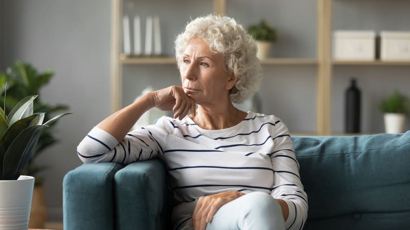 Brooding woman on her couch at home. By Fizkes. A husband left to help his mother and has settled into her home. His wife is tired of being separated by miles. See what “Ask Amy” advises.