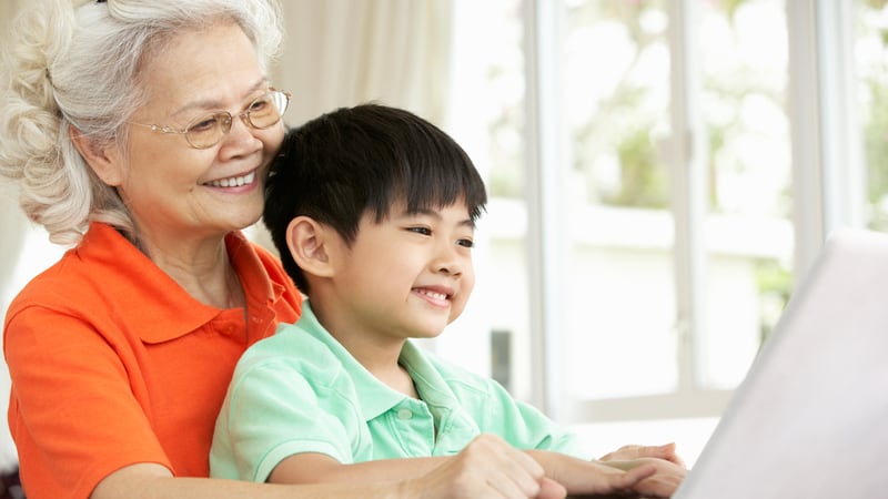An Asian grandmother and her grandson are on a laptop, perhaps playing a puzzle, perhaps the Boomer Jumble puzzles for adults and kids. By Monkey Business Images
