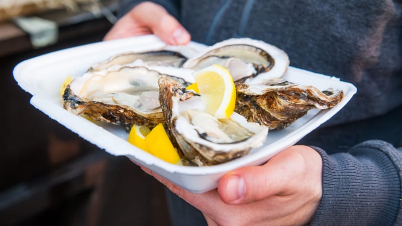 Close up male hand holding take away food tray with fresh opened oysters and lemon slices at street food market, festival, event. Selective focus. copy space