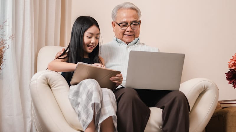 A young girl and her grandfather sit together looking at a tablet and laptop, perhaps working on a puzzle, like the Jumble puzzles - classic and for kids