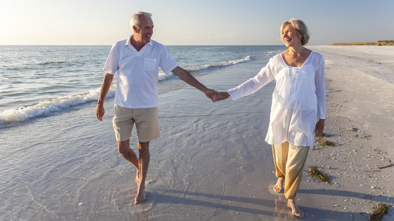 A retired couple walking hand in hand on the beach. By Darren Baker. Article: 6 Things to Consider When Approaching Retirement