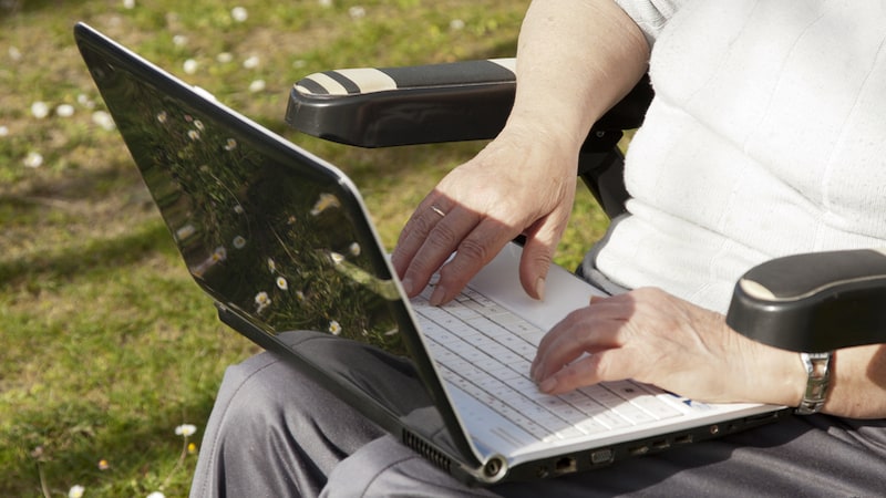 A woman in a wheelchair outside using a laptop, possibly playing a puzzle or other game, like the Boggle six state capitals search. Image by Prudencio Alvarez
