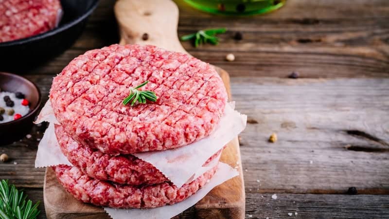 Raw hamburger patties with seasonings such as pepper and herbs sprinkled around. If you want to eat meat but you need to watch your wallet, check out these five cheap cuts of meat.