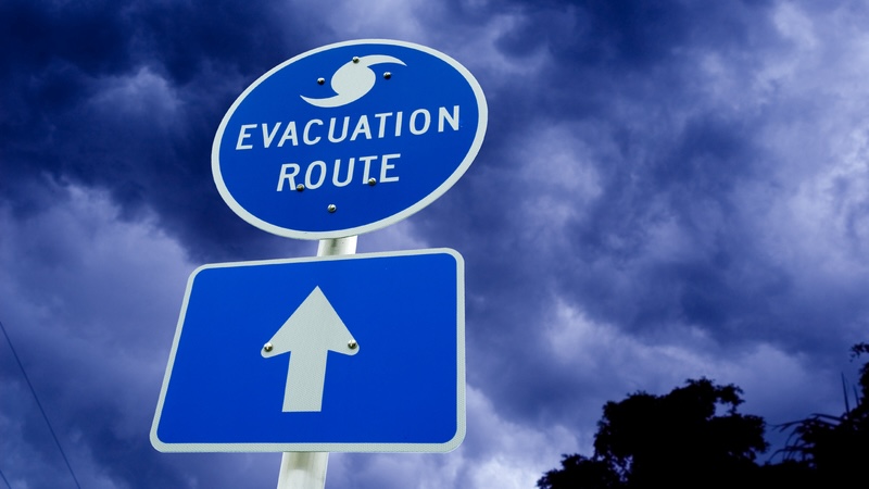 An evacuation route sign with dark storm clouds in the background. For How to Navigate Hurricane Season As You Travel. By Eti Swinford