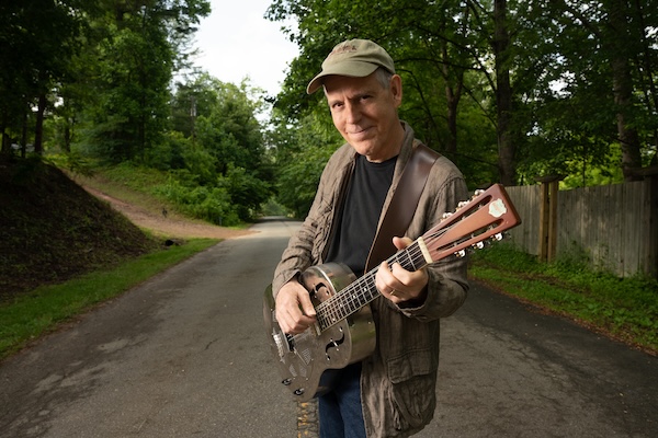 July 17 What's Booming: David Wilcox holding his guitar on a country road. photo credit Lynne Harty 600