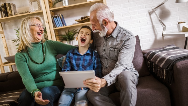 Grandparents and a grandson laughing as they are playing a game on a tablet, like the humorous Jumble puzzles with a songwriter and twins