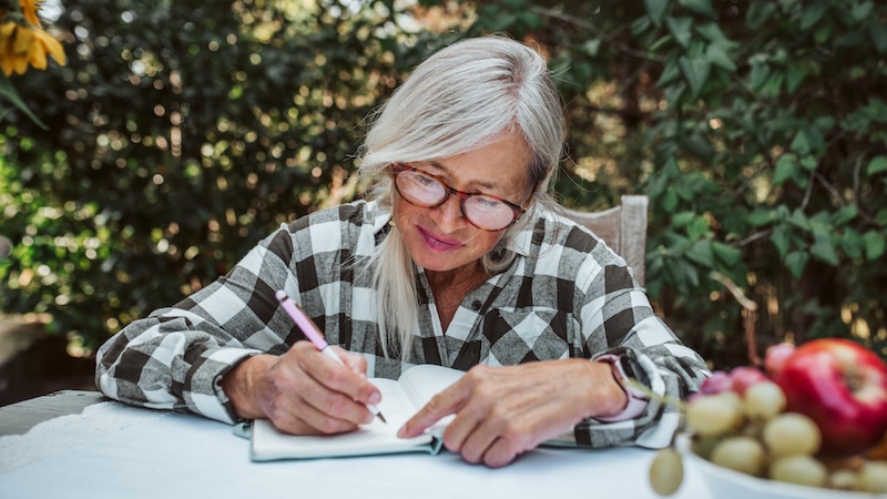 A woman is sitting at a table outside, journaling. Image by Pojoslaw. "Write it down," suggests Mary Derrick Chaney.