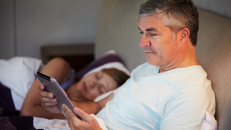 Man using a tablet at bedtime while his wife sleeps. He needs to understand the benefits of minimizing screen time. Image by Monkey Business Images.
