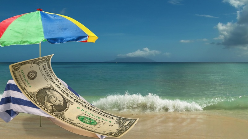 A person-sized dollar bill rests on a beach chair beside the ocean, representing higher summer costs. Banol2007
