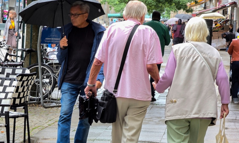 An elderly couple walking slowly on a city street. Their daughter is considering phasing out multigenerational family trips because of their limitations.